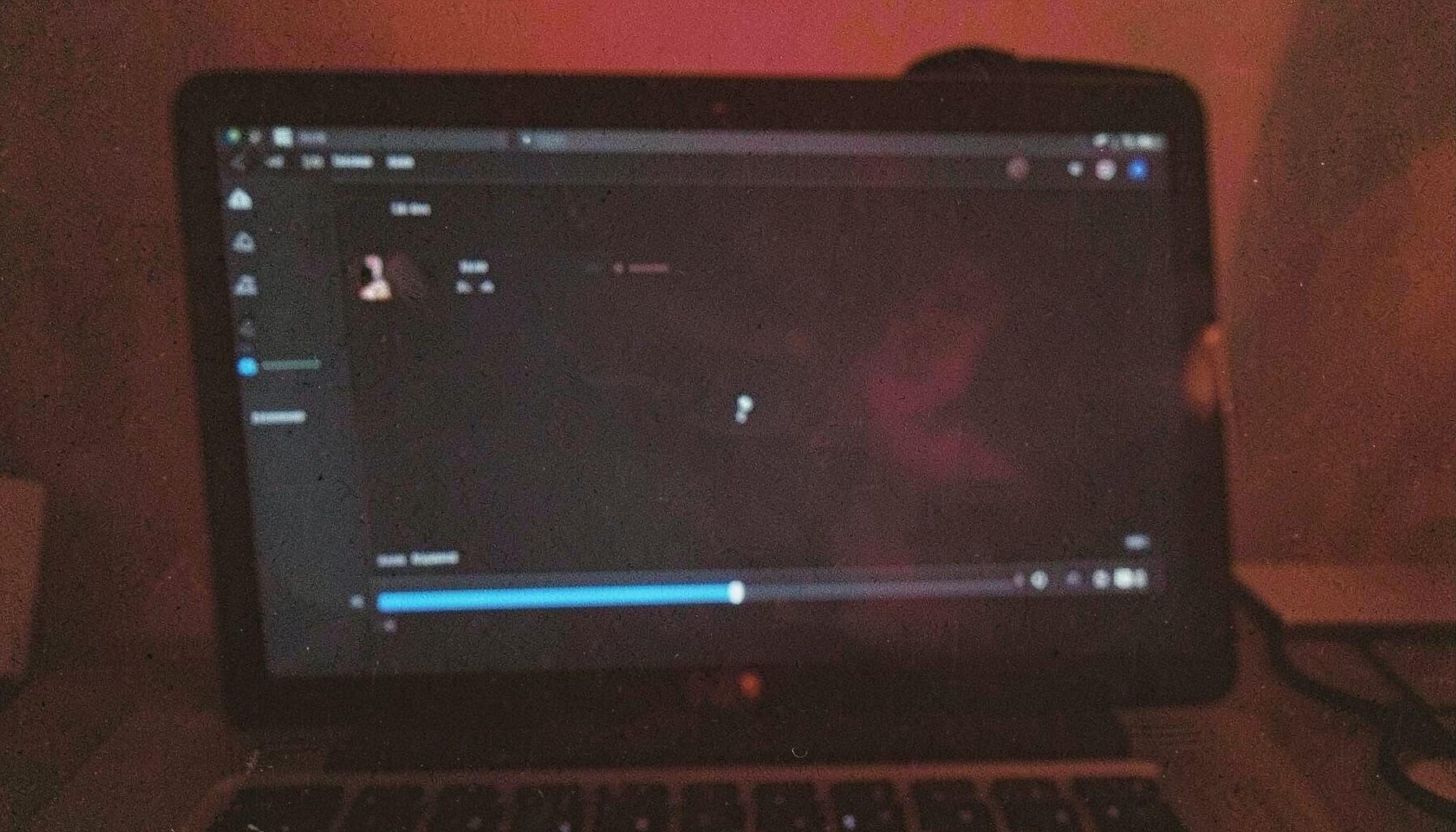 A grainy, low-light photograph of a laptop screen displaying a video player interface with dark UI tones, subtle blue highlights, and a progress bar near the bottom.