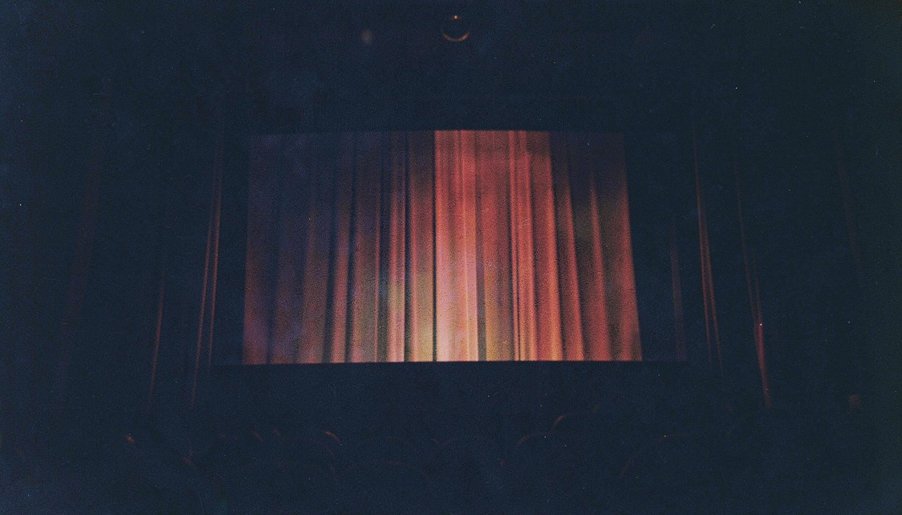 A dark, moody analog film photograph with visible grain showing a cinema screen glow against deep navy shadows, warm amber highlights catching on velvet curtains, evoking the atmosphere of a late-night movie house.