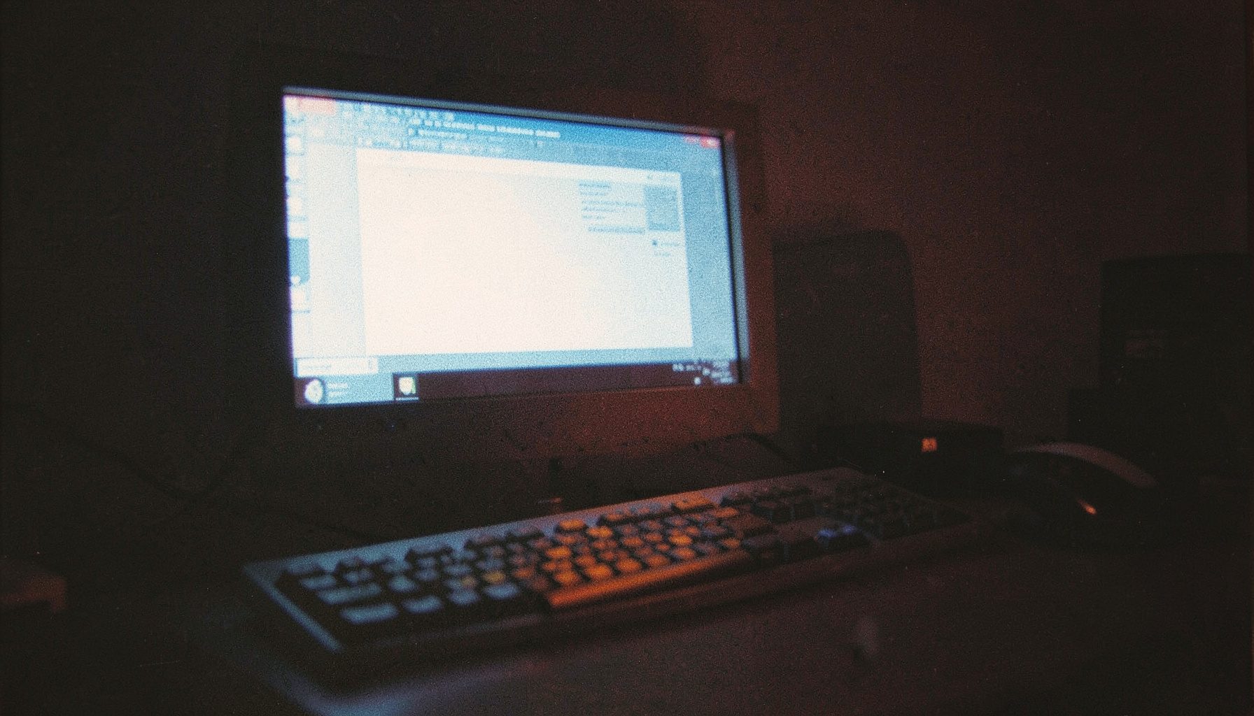 A dim, grainy photograph of a desktop computer screen glowing softly in a dark room, casting cool blue and warm amber tones across a keyboard and mouse.